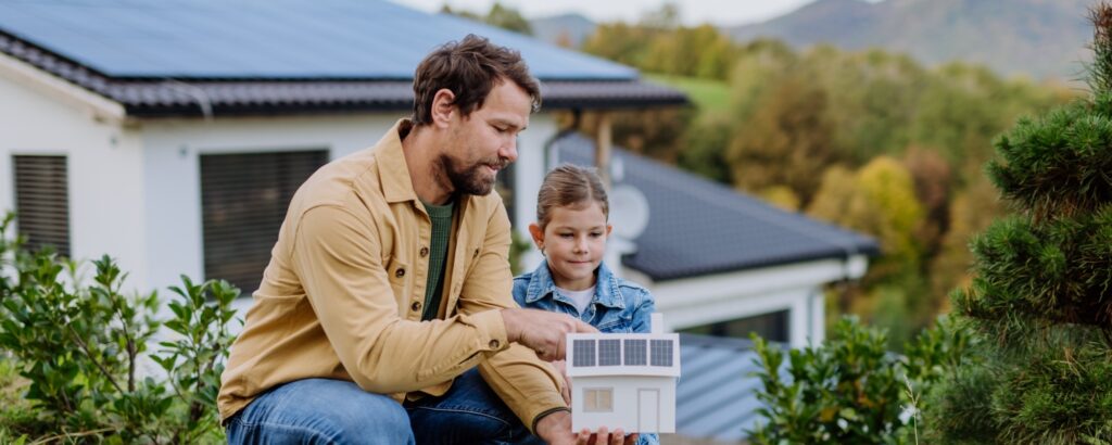 Dekoratives Bild: Vater und Tochter sind im Garten vor ihrem Eigenheim, sie betrachten ein Modellhaus mit Solardeckung.